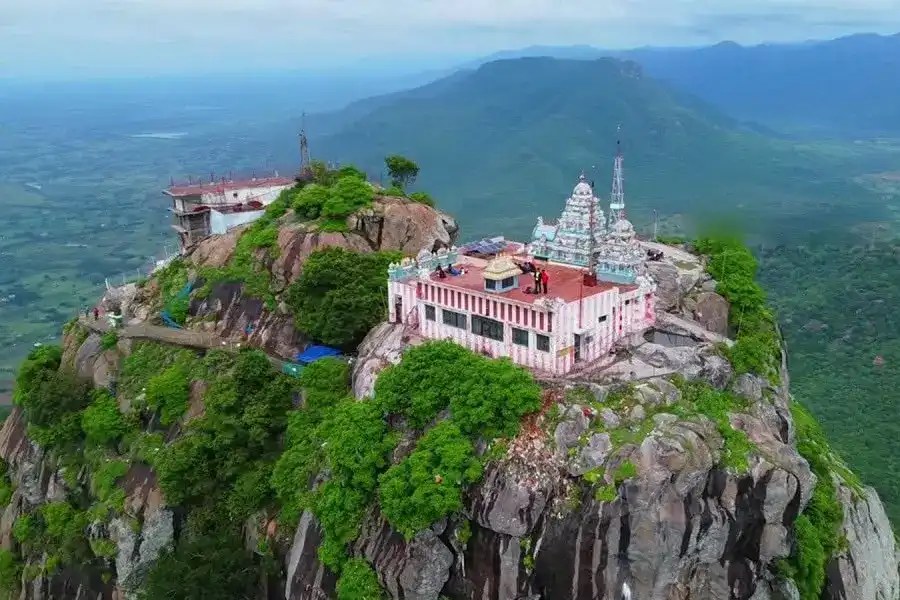 Aerial view of the Mallikarjuna Swamy temple on the rocky summit of Parvathamalai, its pink and white striped sanctum and small gopuram surrounded by green forest and the hills of the Eastern Ghats beyond