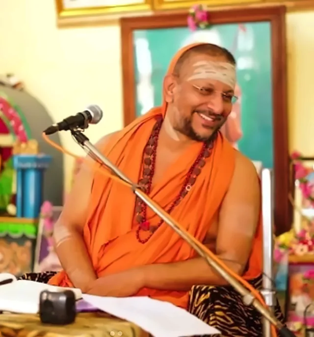 Sri Ramanacharanatirtha (Nochur Swami) smiling during a satsang in Tiruvannamalai, seated at the microphone in saffron robes